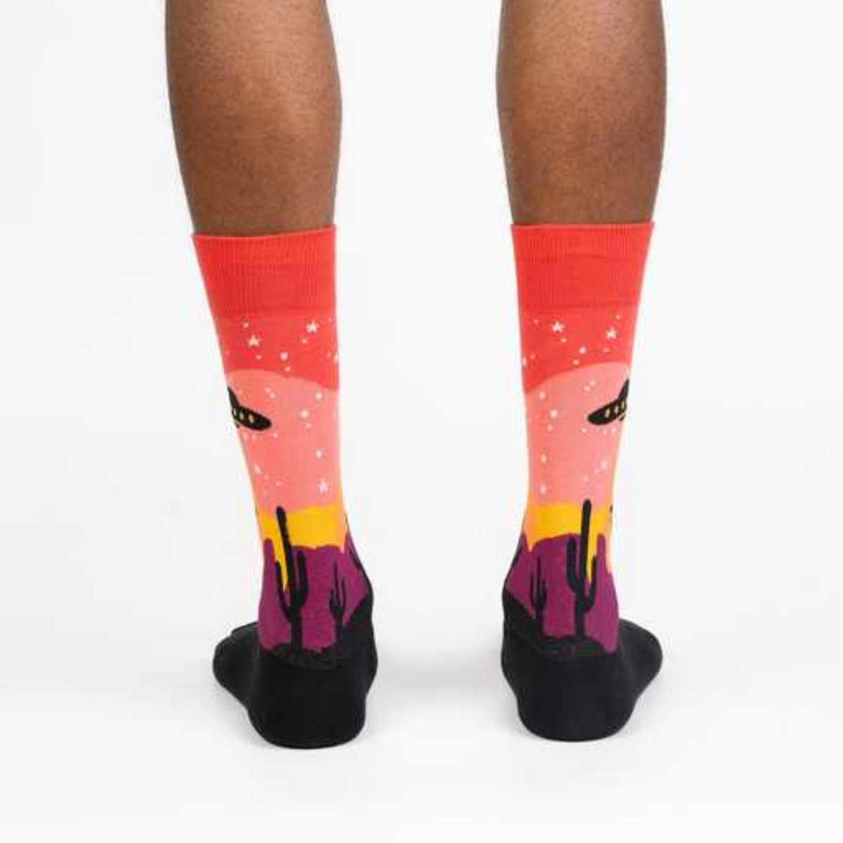 Sock It To Me Area 51 men&#39;s sock  on model from back featuring a sunset with cactus and alien spaceship lifting a cow