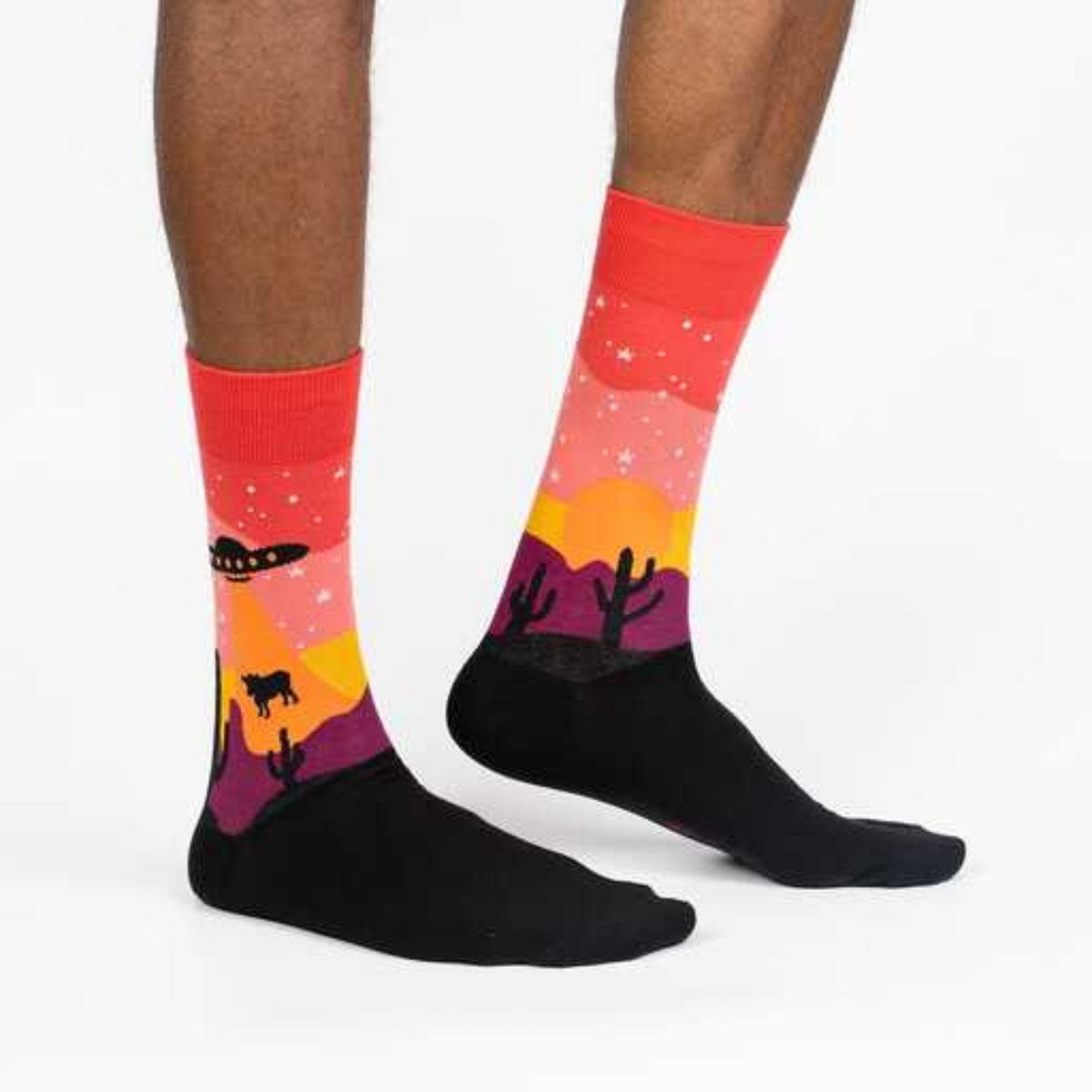 Sock It To Me Area 51 men&#39;s sock  on model from side featuring a sunset with cactus and alien spaceship lifting a cow