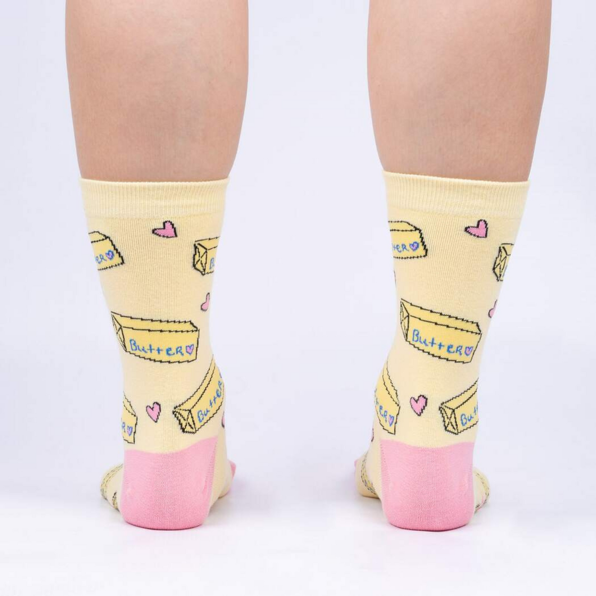 Sock It To Me Butter Me Up women&#39;s yellow crew sock featuring pink hearts, heel and toe with sticks of butter all over on model from behind