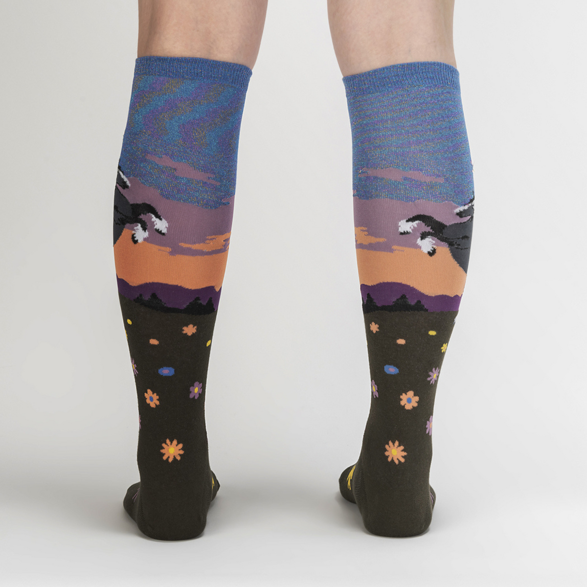 Sock It To Me Black Beauty women&#39;s knee high sock featuring shimmery blue sky with black horse rearing on it&#39;s back legs over a field of flowers worn by model seen from back