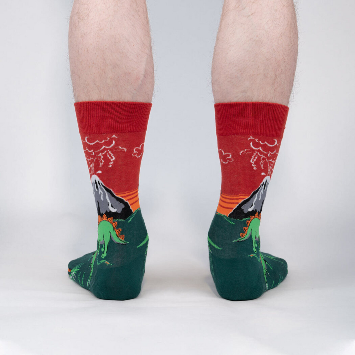 Sock It To Me Dinosaur Days men&#39;s crew socks featuring red and green socks with glow in the dark exploding volcanoes and dinosaurs. Socks shown on model from behind. 