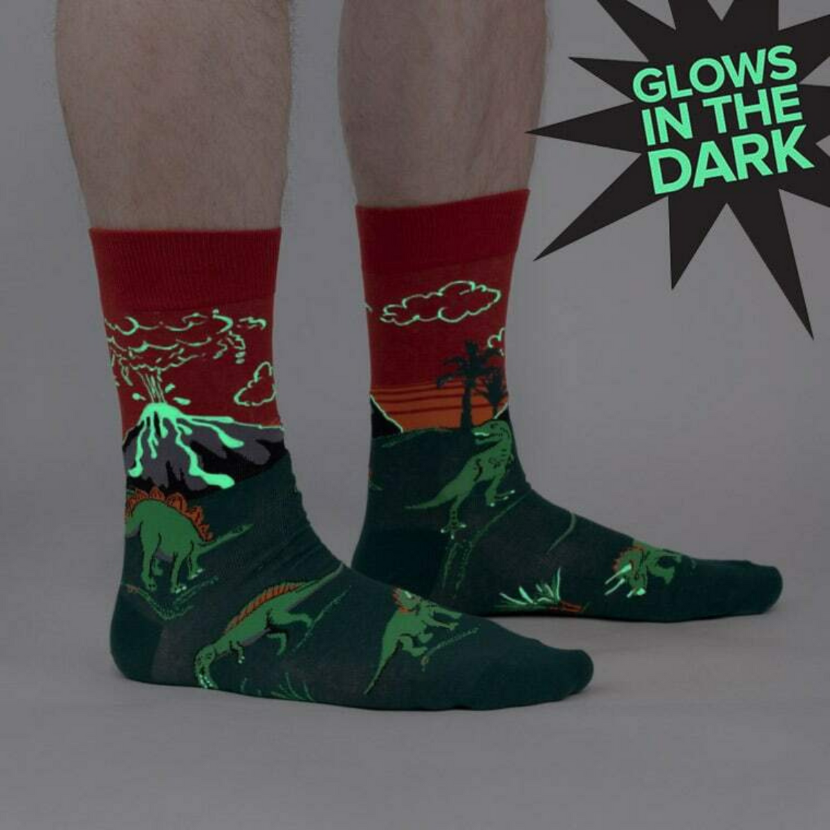 Sock It To Me Dinosaur Days men&#39;s crew socks featuring red and green socks with glow in the dark exploding volcanoes and dinosaurs. Socks shown on model from the side. 