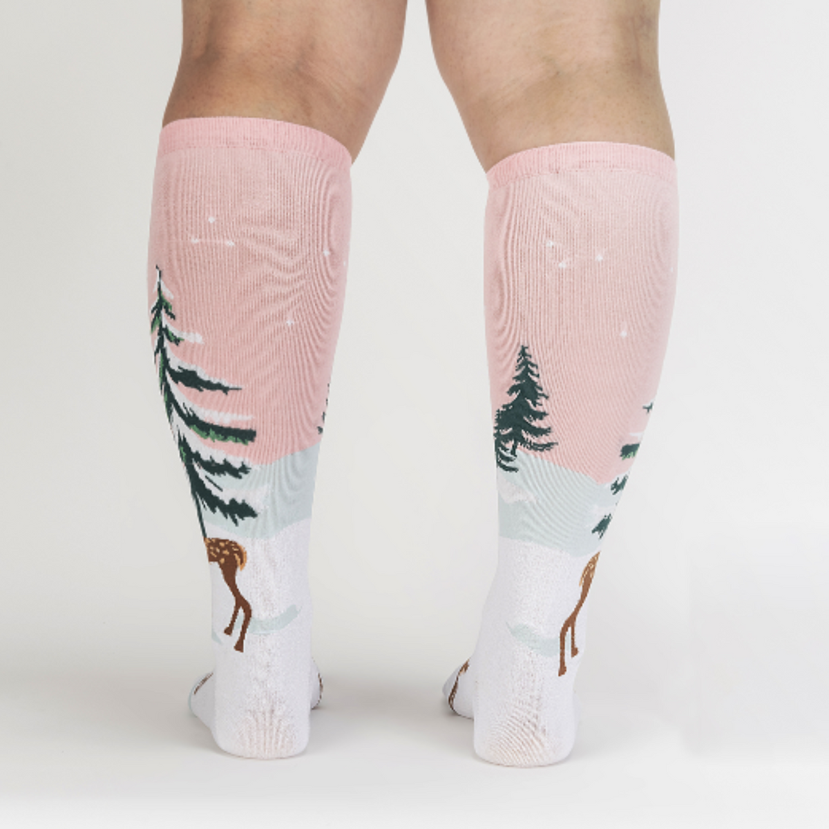 Sock It To Me Doe-nt Forget Your Scarf women&#39;s knee high sock featuring pink sock with snow-covered fir tree and deer with pink scarf. Socks worn by model seen from behind. 