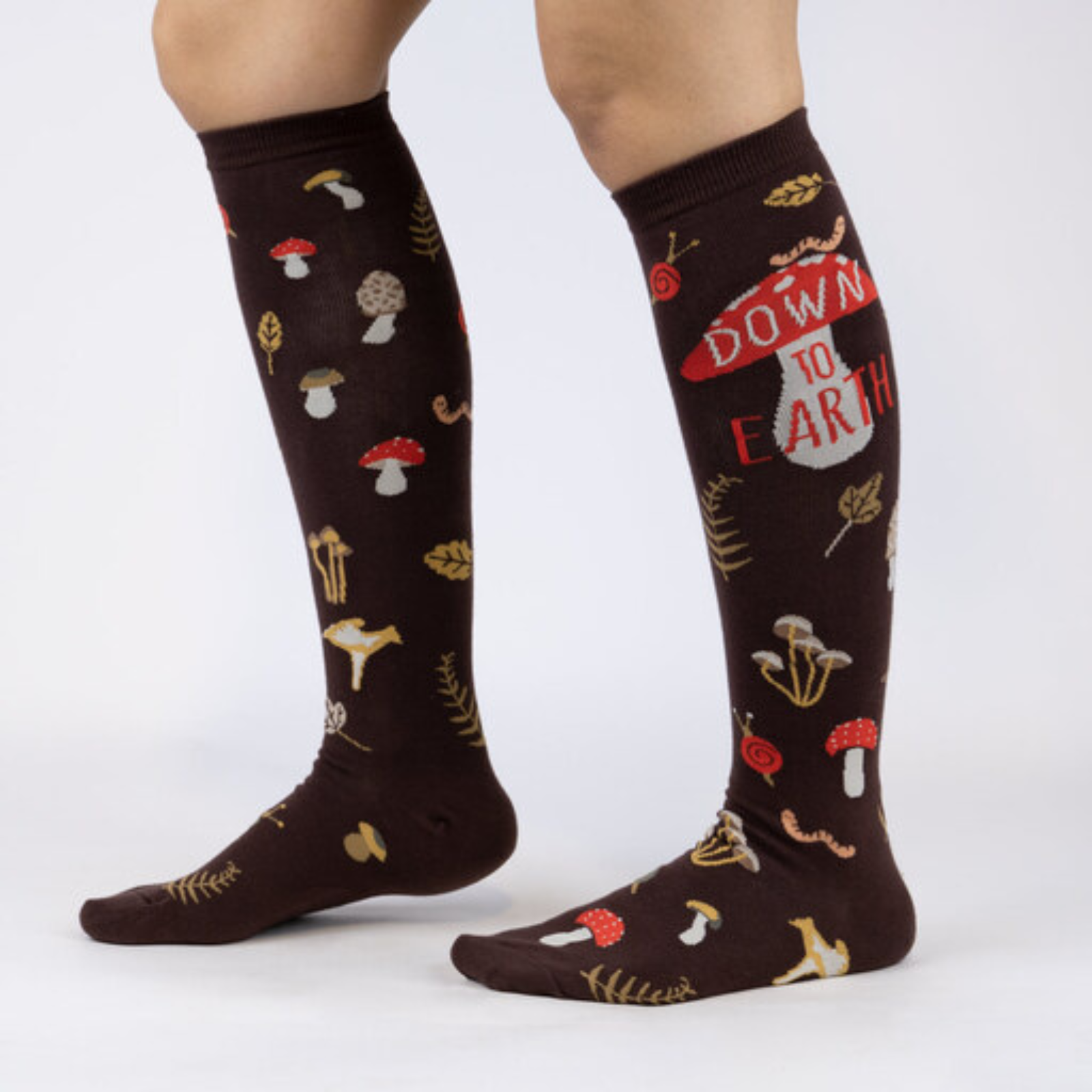 Sock It To Me Down to Earth women's knee high sock featuring brown sock with different mushrooms all over and the phrase "Down to Earth". Socks worn by model seen from side. 