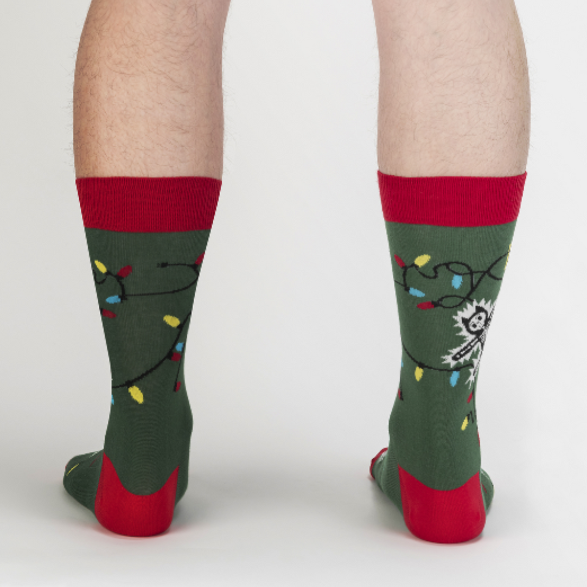 Sock It To Me Eating Light This Holiday (GLOWS IN THE DARK) women&#39;s and men&#39;s crew socks featuring green sock with red cuff, heel, and toe with cartoon cat being electrocuted by Christmas lights. Socks worn by male model seen from back. 