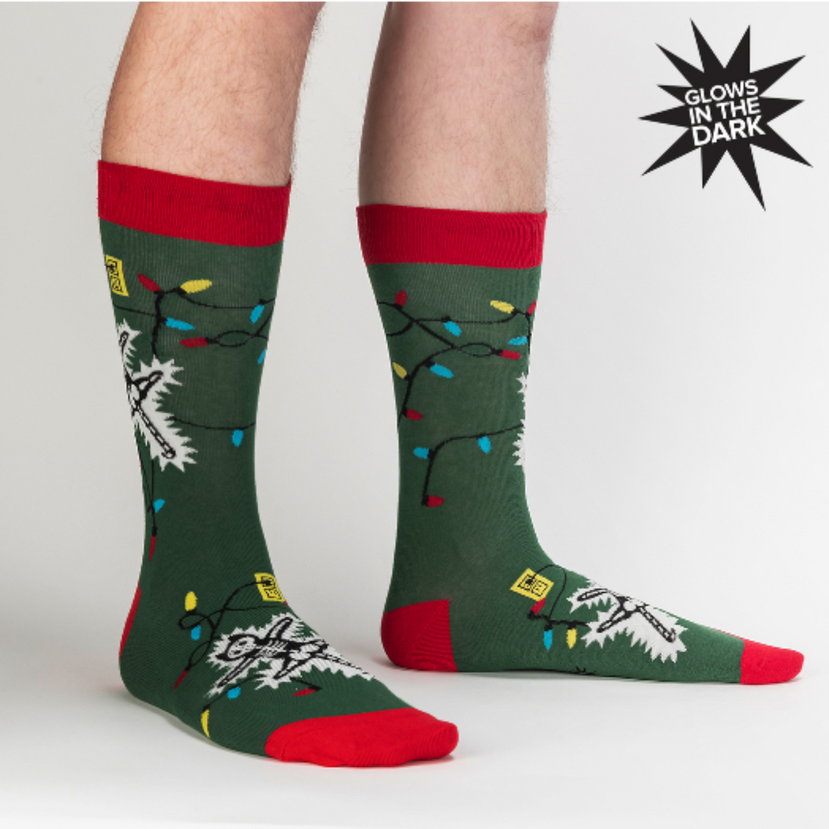 Sock It To Me Eating Light This Holiday (GLOWS IN THE DARK) women&#39;s and men&#39;s crew socks featuring green sock with red cuff, heel, and toe with cartoon cat being electrocuted by Christmas lights. Socks worn by male model seen from side. 