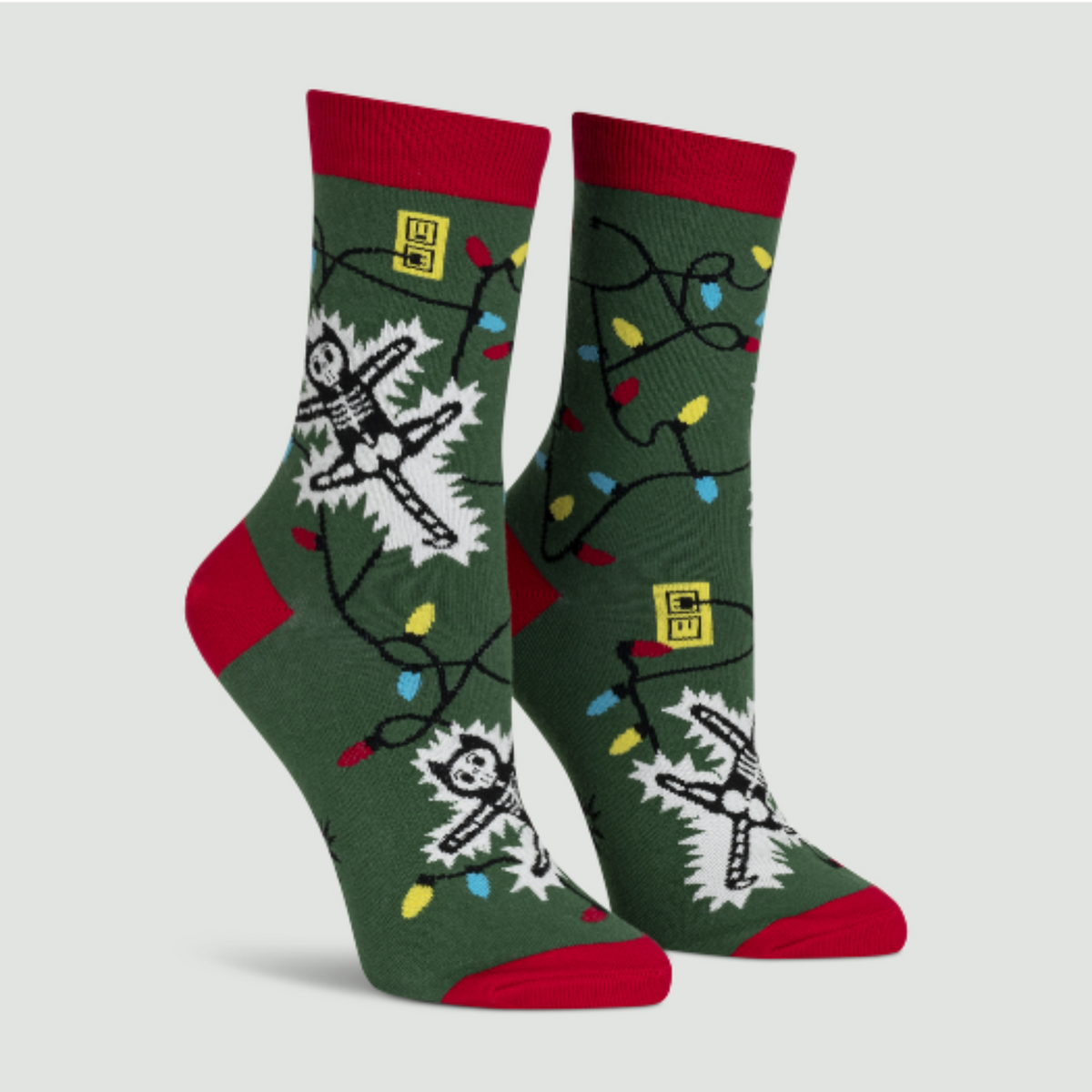 Sock It To Me Eating Light This Holiday (GLOWS IN THE DARK) women&#39;s and men&#39;s crew socks featuring green sock with red cuff, heel, and toe with cartoon cat being electrocuted by Christmas lights. Socks shown on display feet. 