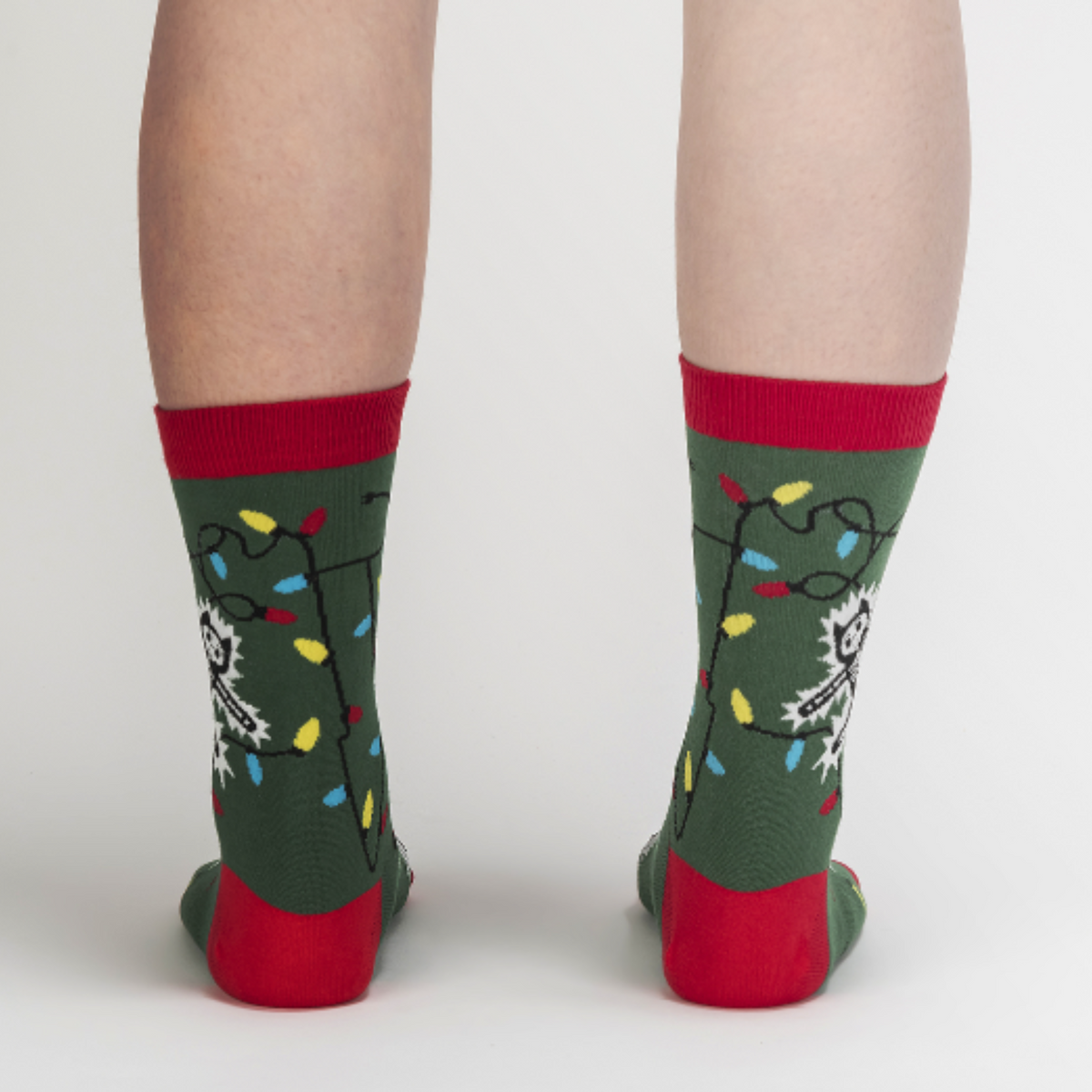 Sock It To Me Eating Light This Holiday (GLOWS IN THE DARK) women&#39;s and men&#39;s crew socks featuring green sock with red cuff, heel, and toe with cartoon cat being electrocuted by Christmas lights. Socks worn by female model seen from back.. 