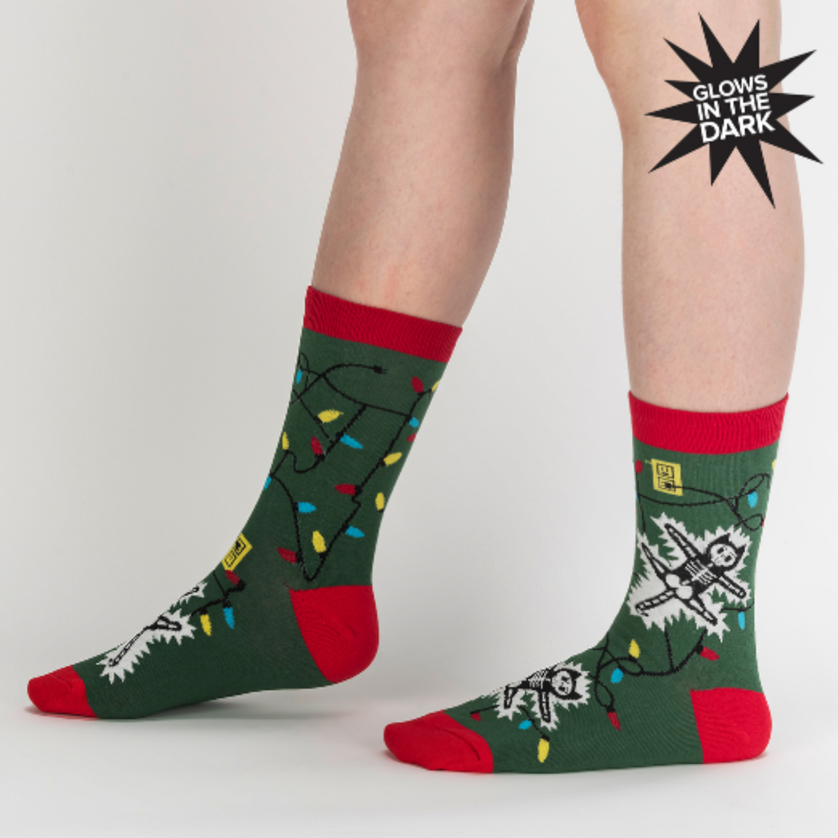 Sock It To Me Eating Light This Holiday (GLOWS IN THE DARK) women&#39;s and men&#39;s crew socks featuring green sock with red cuff, heel, and toe with cartoon cat being electrocuted by Christmas lights. Socks worn by female model seen from side. 