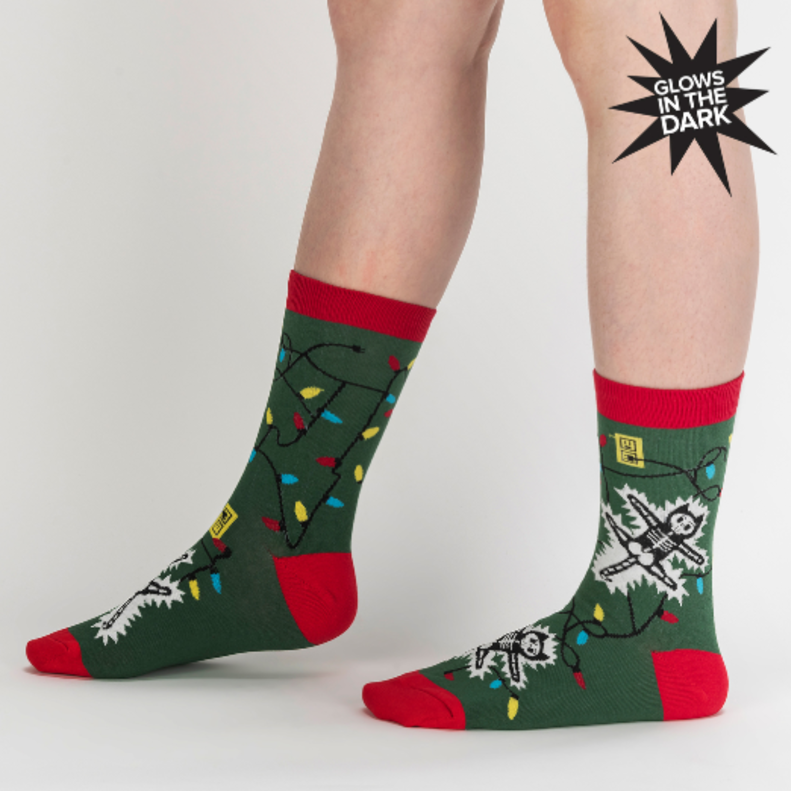Sock It To Me Eating Light This Holiday (GLOWS IN THE DARK) women's and men's crew socks featuring green sock with red cuff, heel, and toe with cartoon cat being electrocuted by Christmas lights. Socks worn by female model seen from side. 