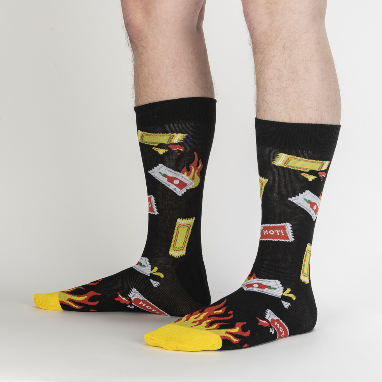 Sock It To Me Extra Hot men's sock featuring black crew sock with hot sauce packets and flames at toes worn by model seen from side