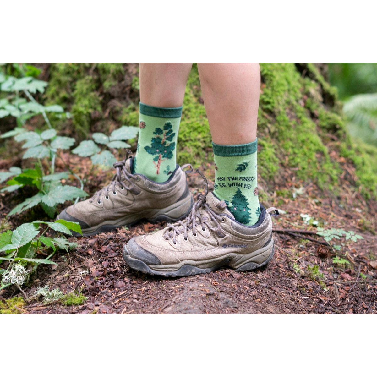 Sock It To Me May the Forest Be With You women&#39;s crew sock featuring a green sock with trees and pinecones and &quot;May the forest be with you&quot; written. Socks worn by a model outdoors. 