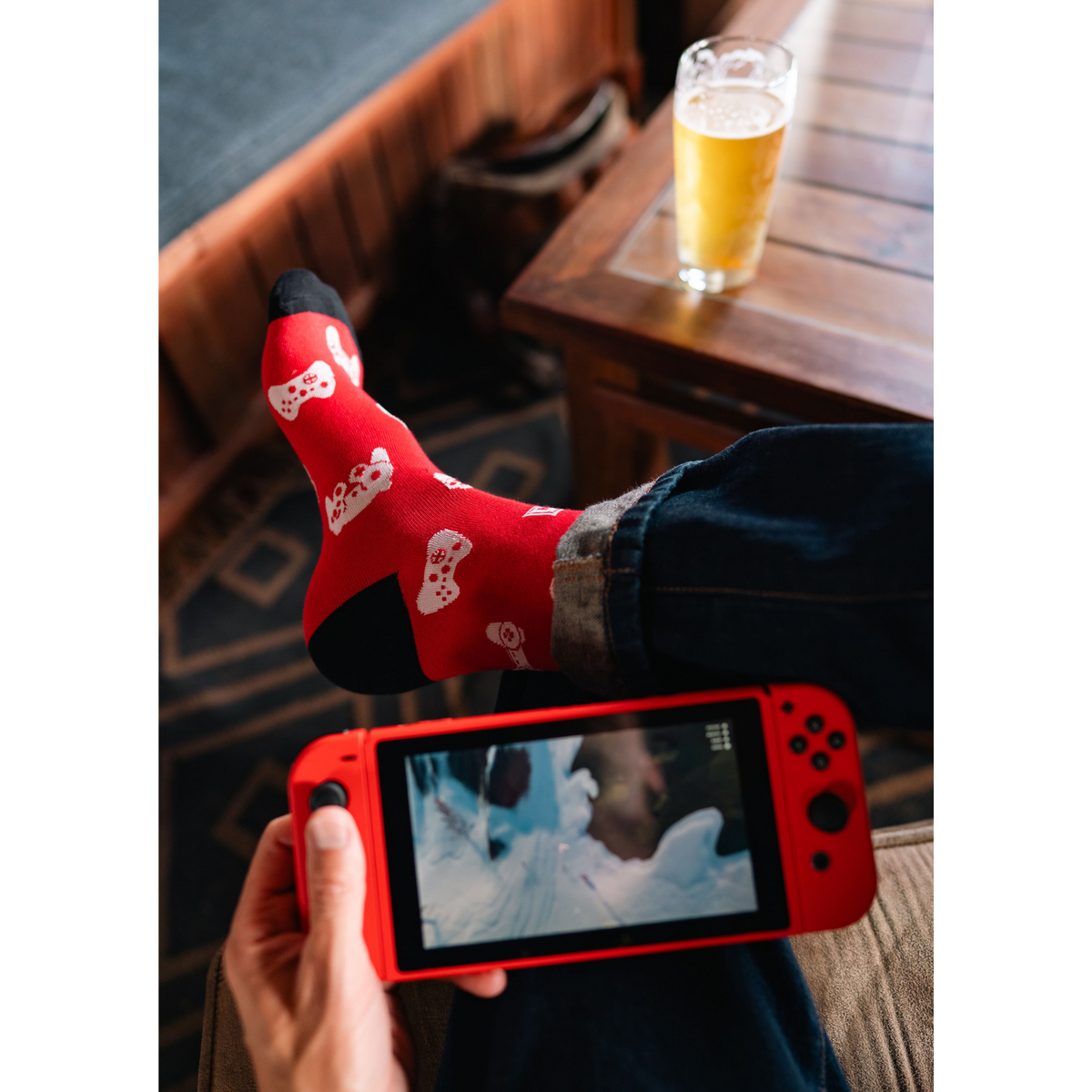 Sock It To Me Multi-Player men&#39;s crew sock featuring red sock with black cuff, heel, and toes and white video game controllers all over. Socks shown on model in Mendocino playing video game while drinking a beer. 