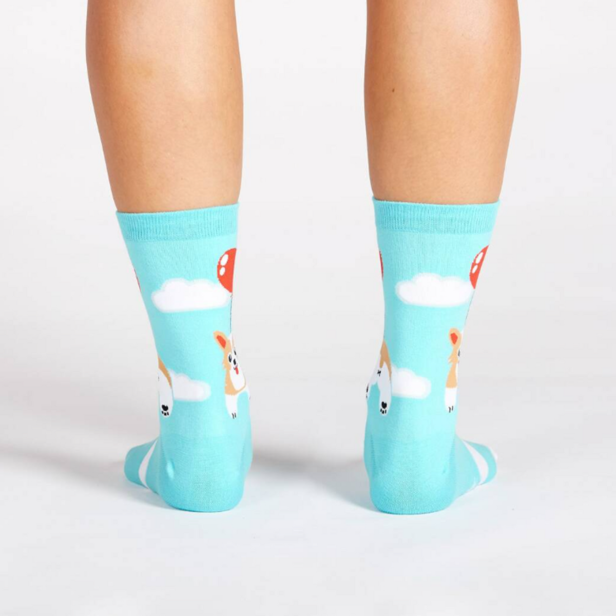 Sock It To Me Pup, Pup and Away women&#39;s crew socks featuring light blue socks with clouds all over and a Corgi dog floating by a red balloon, socks show front and back of Corgi dog. Socks worn by model seen from back. 