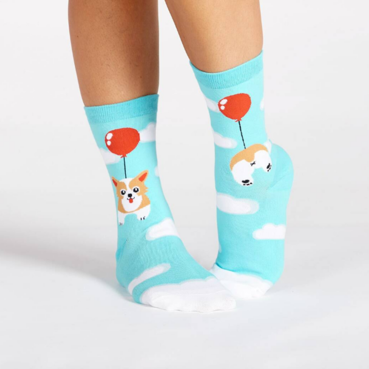 Sock It To Me Pup, Pup and Away women&#39;s crew socks featuring light blue socks with clouds all over and a Corgi dog floating by a red balloon, socks show front and back of Corgi dog. Socks worn by model seen from side.