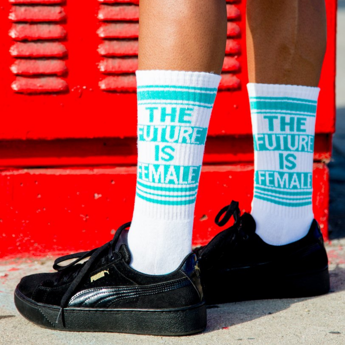 foot model in black shoes wearing Gumball Poodle Future is Female women&#39;s and men&#39;s sock featuring a white sock with teal writing that says the Future is Female in all caps