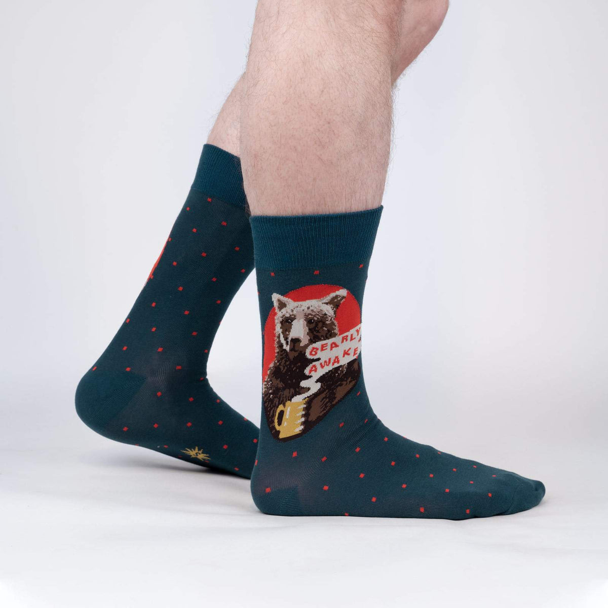 Sock It To Me Bearly Awake women&#39;s and men&#39;s crew sock featuring blue sock with small red polka dots and picture of bear holding a cup of coffee and the saying &quot;bearly awake&quot;. Socks worn by model seen from side.