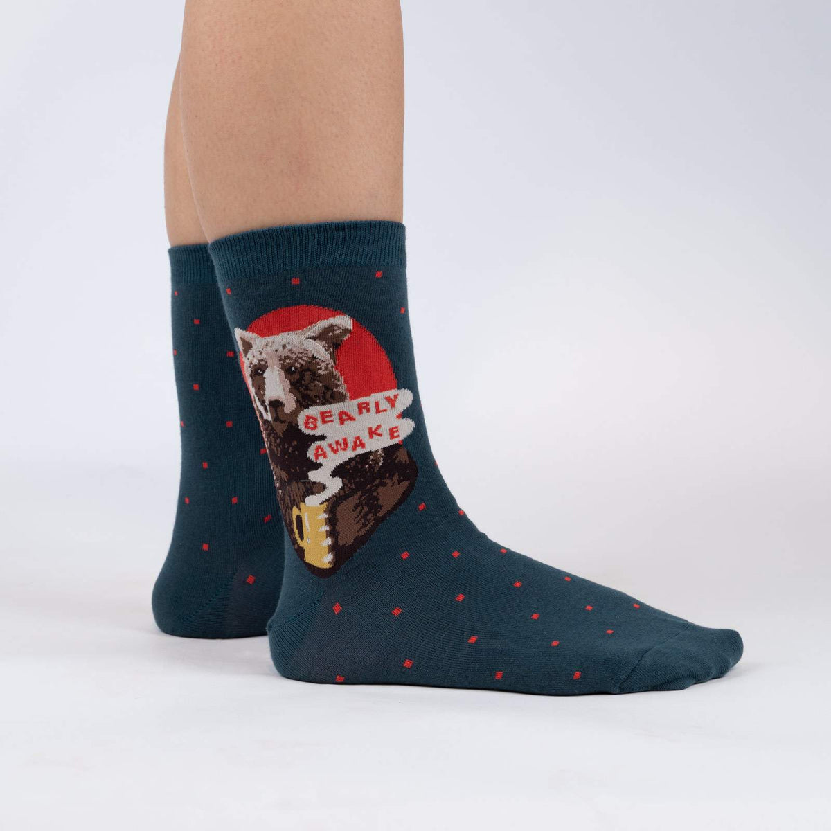 Sock It To Me Bearly Awake women&#39;s and men&#39;s crew sock featuring blue sock with small red polka dots and picture of bear holding a cup of coffee and the saying &quot;bearly awake&quot;. Socks worn by model seen from side.