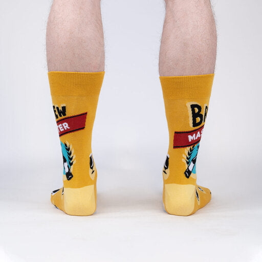 Sock It To Me Brew Master men&#39;s crew sock. Featuring yellow sock with &quot;Brew Master&quot; and various beer glasses all over. Socks worn by model seen from behind.