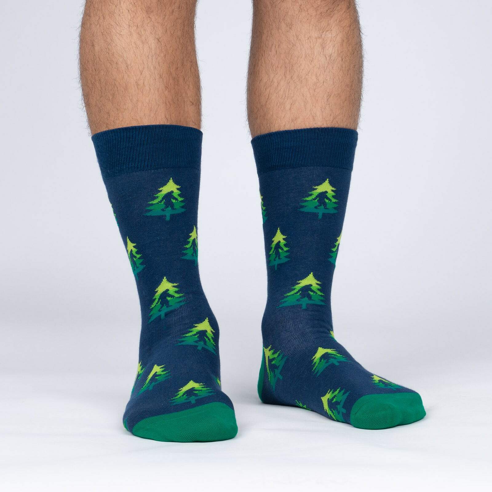 Sock It To Me Do You Tree What I Tree men's crew sock featuring blue sock with green trees that have a Sasquatch silhouette inside them. Socks worn by model seen from the front. 