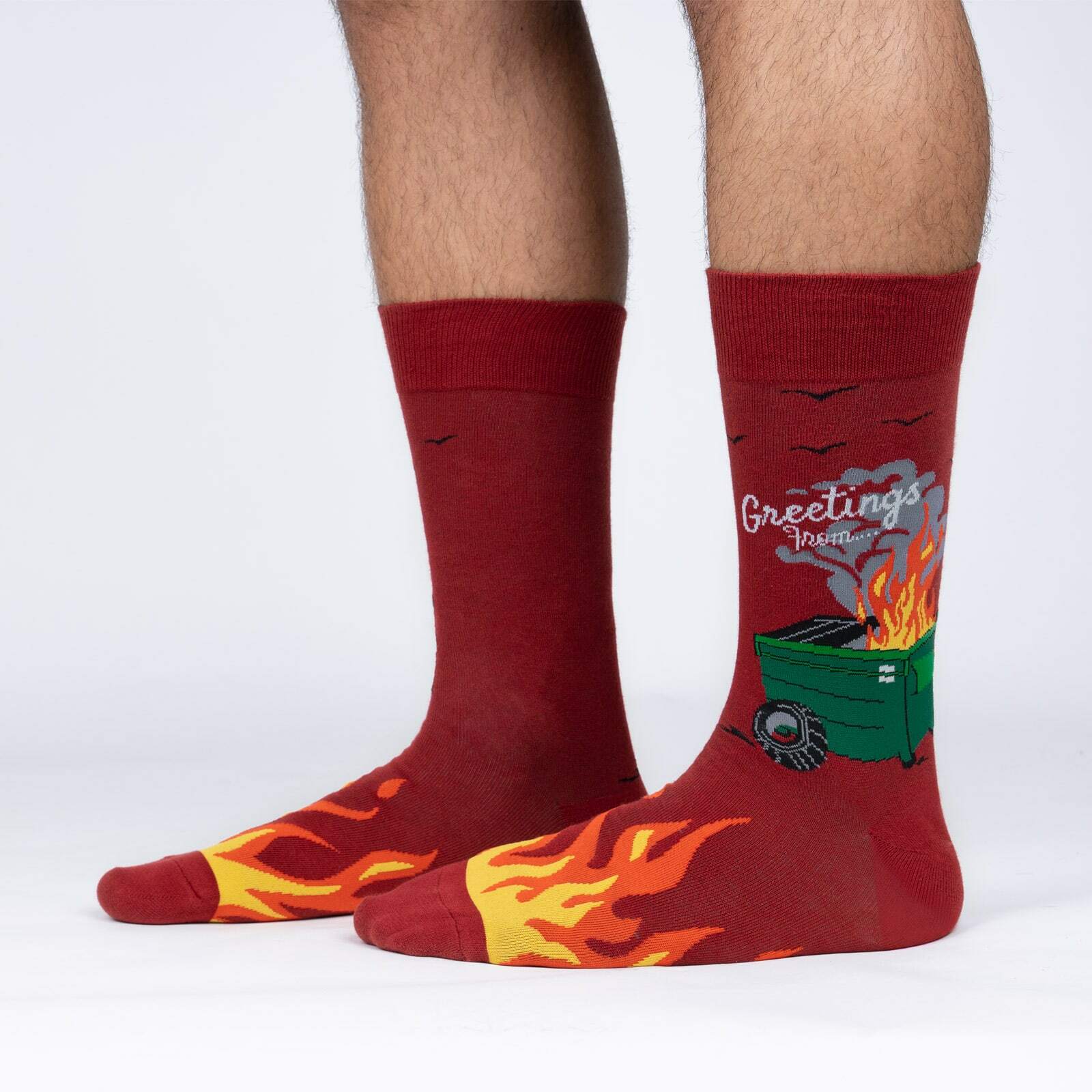 Sock It To Me Dumpster Fire men's crew sock featuring red sock with the words "Greetings form ..." over a green garbage dumpster on fire. Socks worn by model seen from the side. 