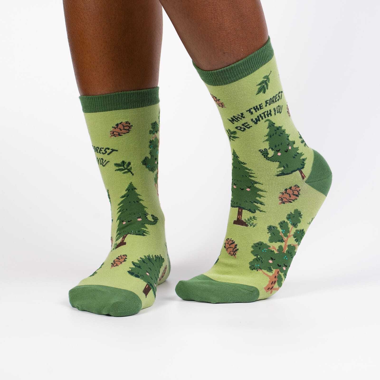 Sock It To Me May the Forest Be With You women's crew sock featuring a green sock with trees and pinecones and "May the forest be with you" written. Socks worn by model seen from the side. 