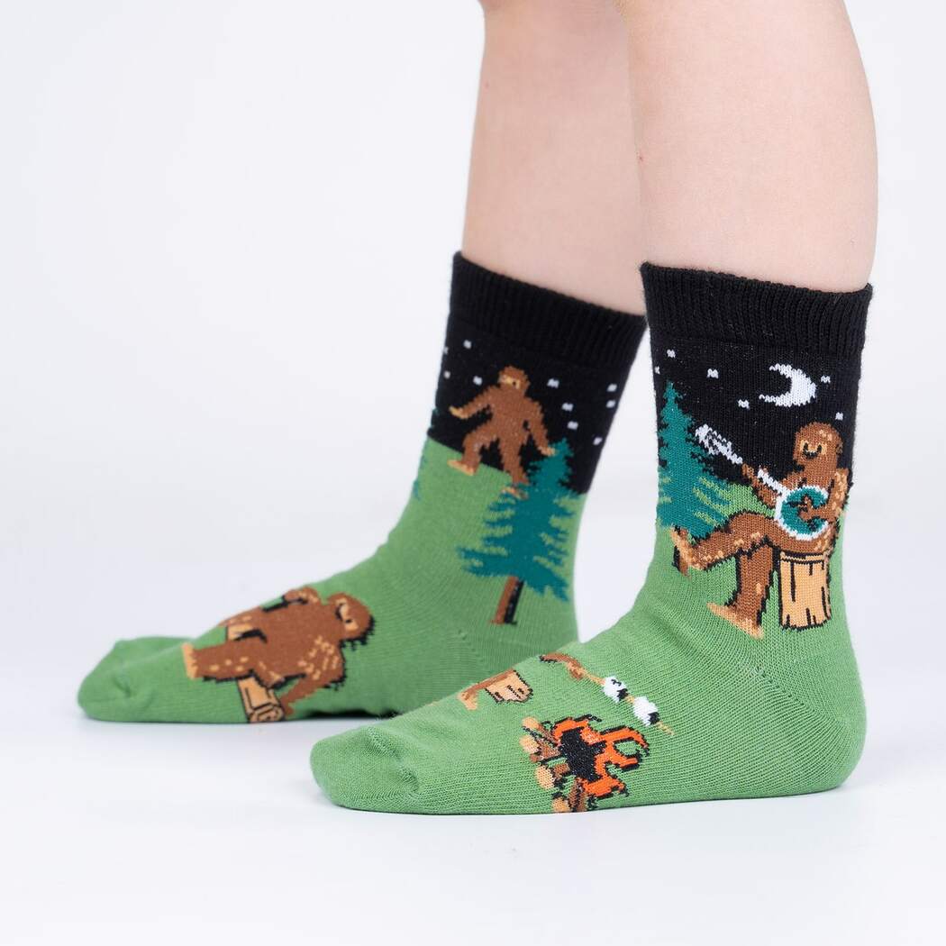 Sock It To Me Sasquatch Camp Out 3-pack kids&#39; crew socks featuring Sasquatch camping and playing the banjo socks on model seen from side. 