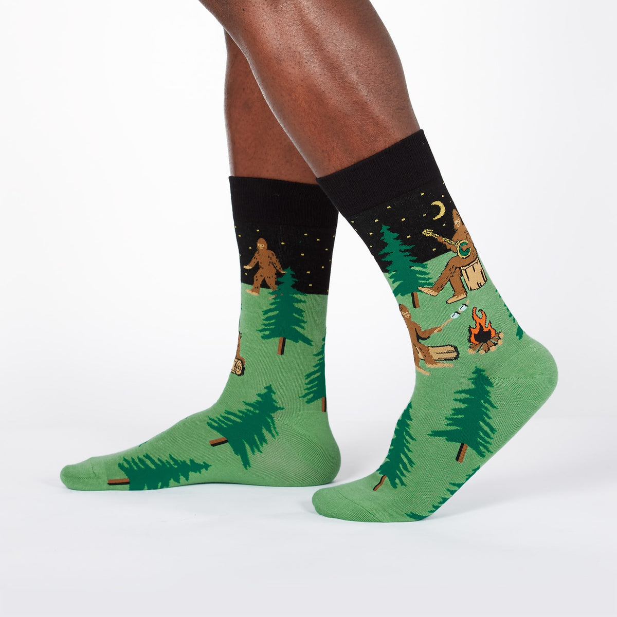 Sock It To Me Sasquatch Camp Out men&#39;s socks on model showing Sasquatch playing a big banjo and little Sasquatch roasting marshmallows by a campfire
